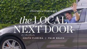 A smiling man in a blue shirt waves from the driver’s seat of a gray car. Overlaid text reads: Corcoran presents The Local Next Door, South Florida | Palm Beach. Green foliage is visible in the background.