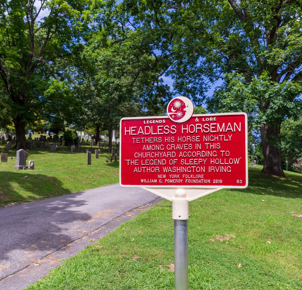 A red rectangular sign with white text in a graveyard tells of the Headless Horseman from a story by Washington Irving.