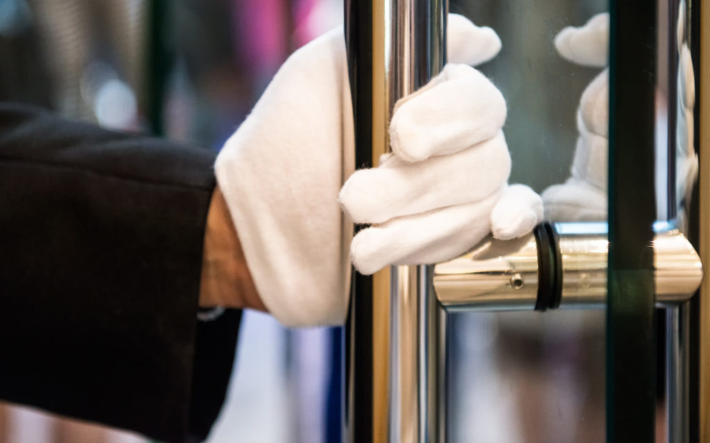 doorman_thumb A person wearing a black sleeve and white gloves firmly grips the handle of a glass door, suggesting a professional or service role such as a doorman or concierge.