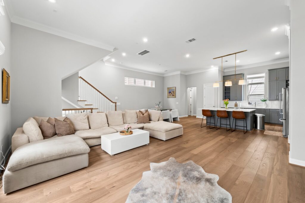 Spacious modern living room, featuring a large beige sectional sofa, white coffee table, wood flooring, and an open kitchen with gray cabinets, breakfast bar, and pendant lights.
