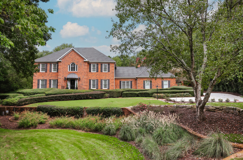 132 Branford Place, Athens A large two-story brick house with white trim and multiple windows sits behind manicured lawns, shrubs, and trees under a partly cloudy sky in a picturesque college town.