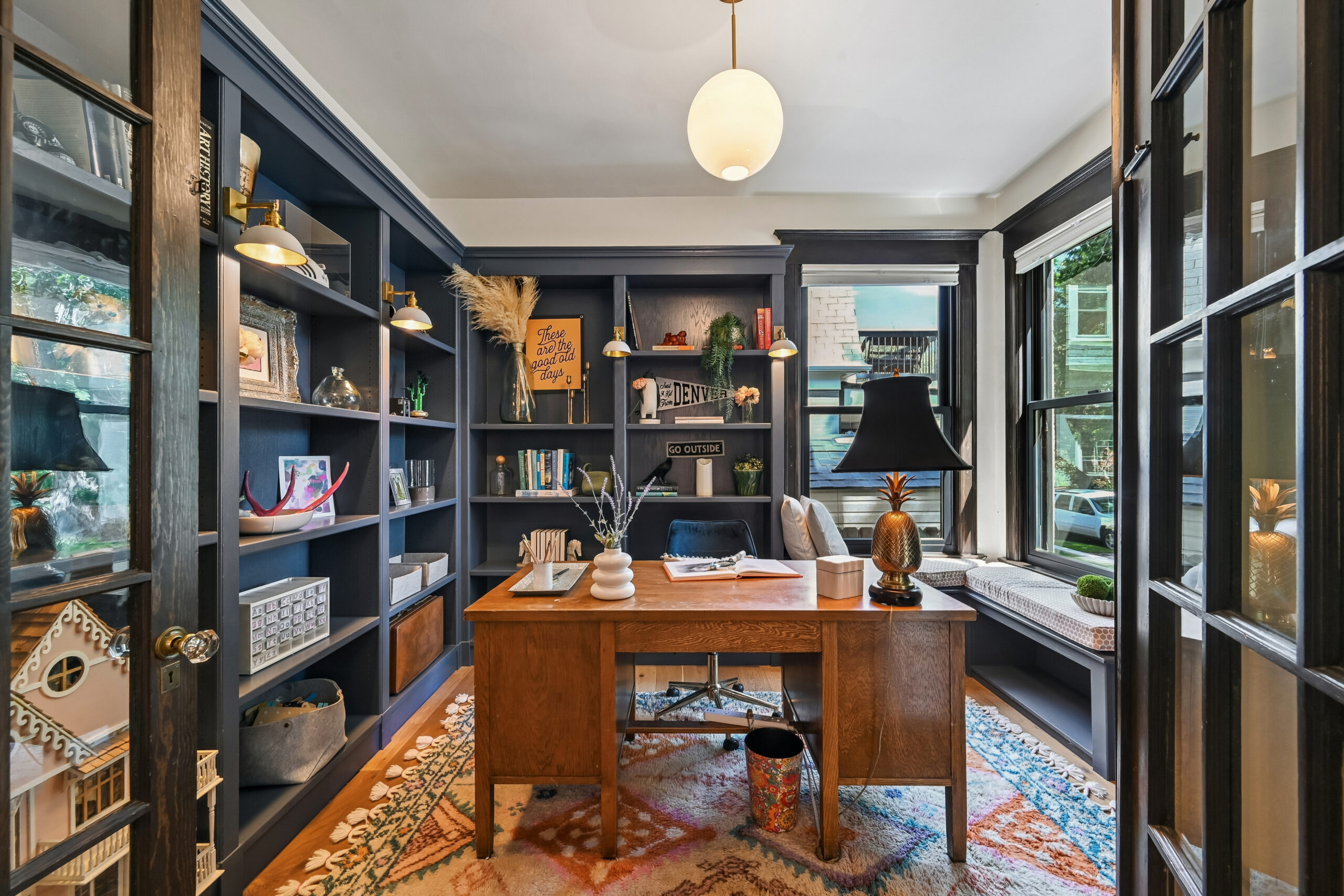 1539 Saint Paul Street Home office featuring a wooden desk, a pineapple-shaped lamp, and dark blue built-in bookshelves filled with books and decor, all situated on a colorful patterned rug.