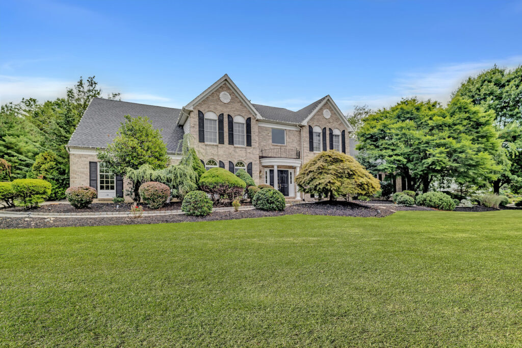 A large, two-story brick house with black shutters and arched windows sits in a vibrant college town, surrounded by manicured bushes, trees, and a spacious green lawn under a clear blue sky.