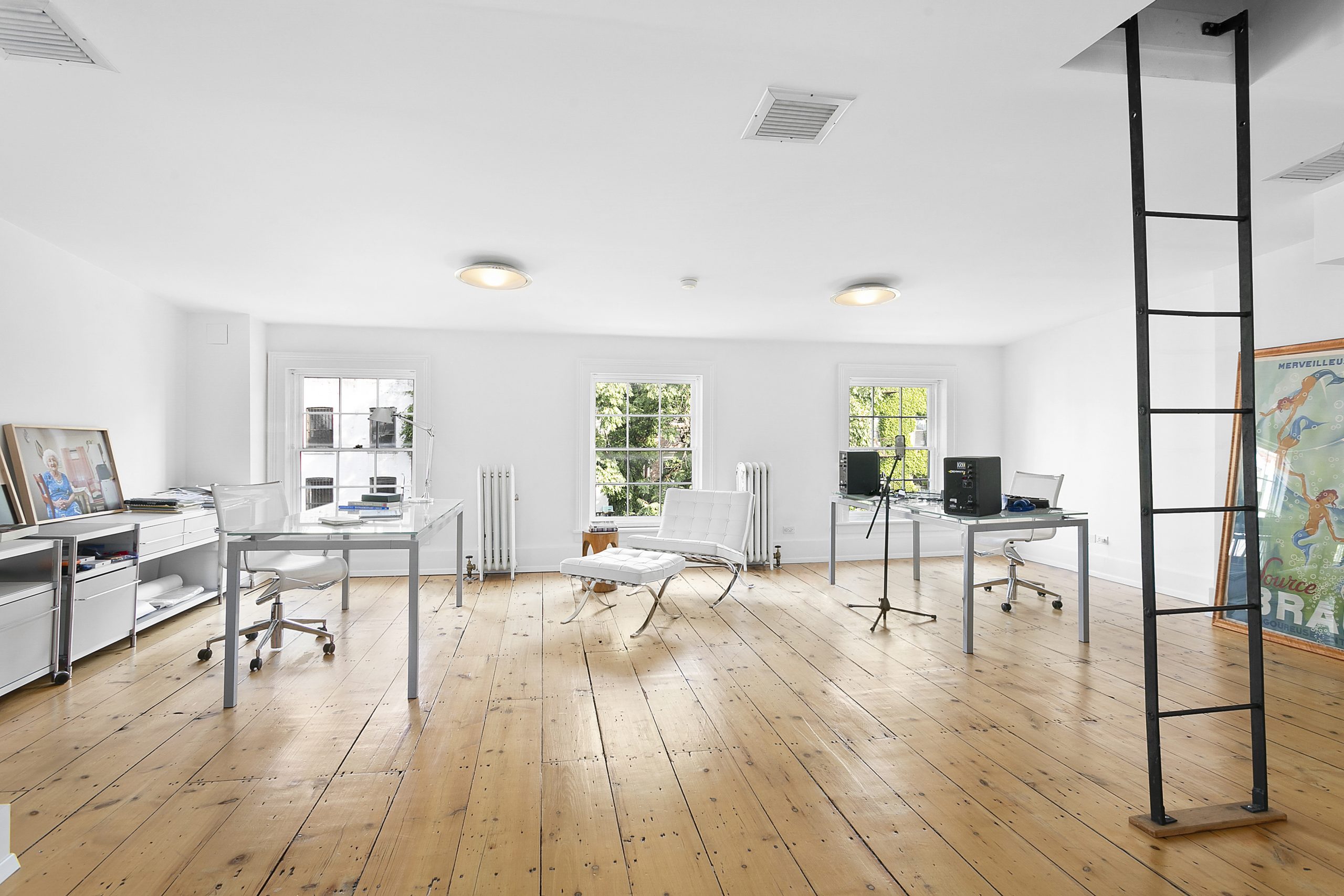 Bright, modern office space ideal for home offices, featuring large windows, wooden floors, white walls, minimal furniture including desks, chairs, computers, framed artwork, and a black metal ladder leading to an upper level.