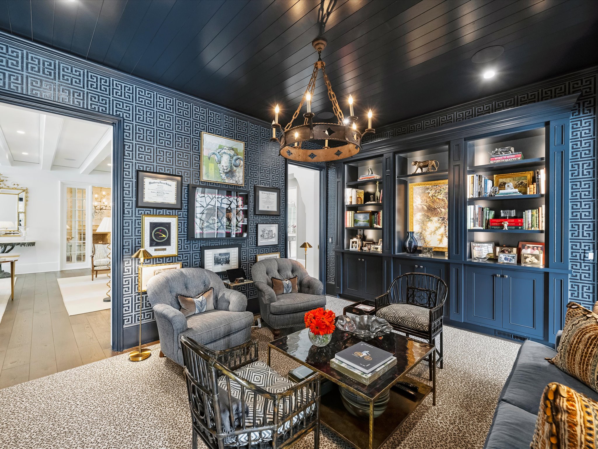 2414 Forest Drive A sitting area featuring two gray armchairs, two patterned black wooden chairs, and a glass-topped coffee table, set against navy blue geometric wallpaper and built-in bookshelves.