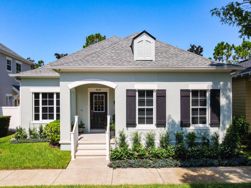 A single-story house with light-colored walls, dark shutters, a small front porch with white railings, neatly trimmed shrubs, and a manicured lawn.