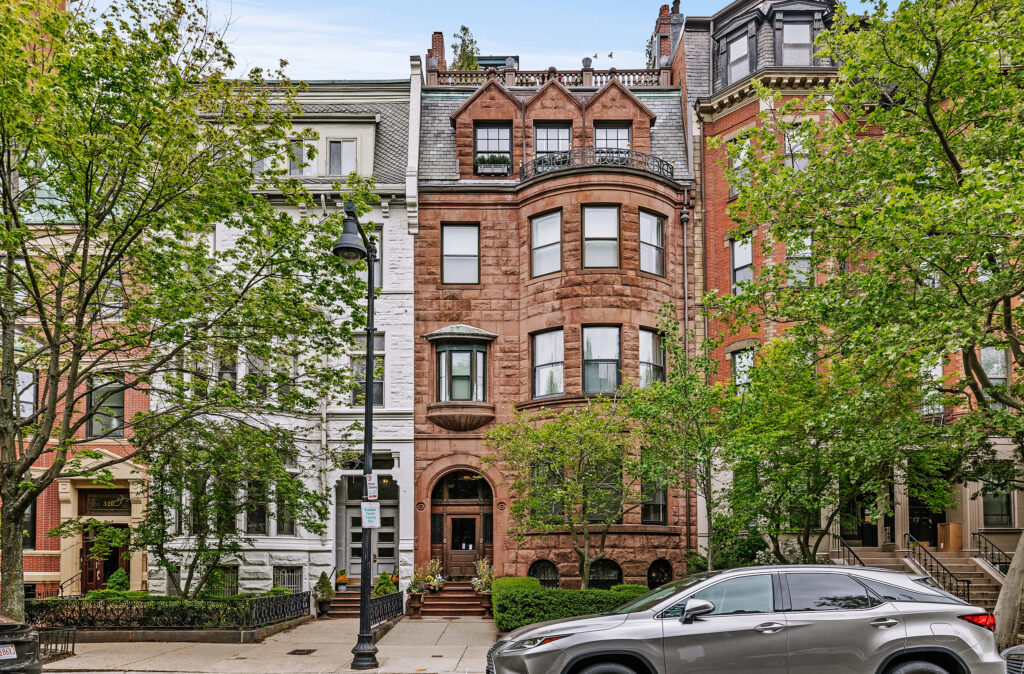 A historic brownstone townhouse with arched windows and a detailed facade sits between two other ornate buildings on a tree-lined street.