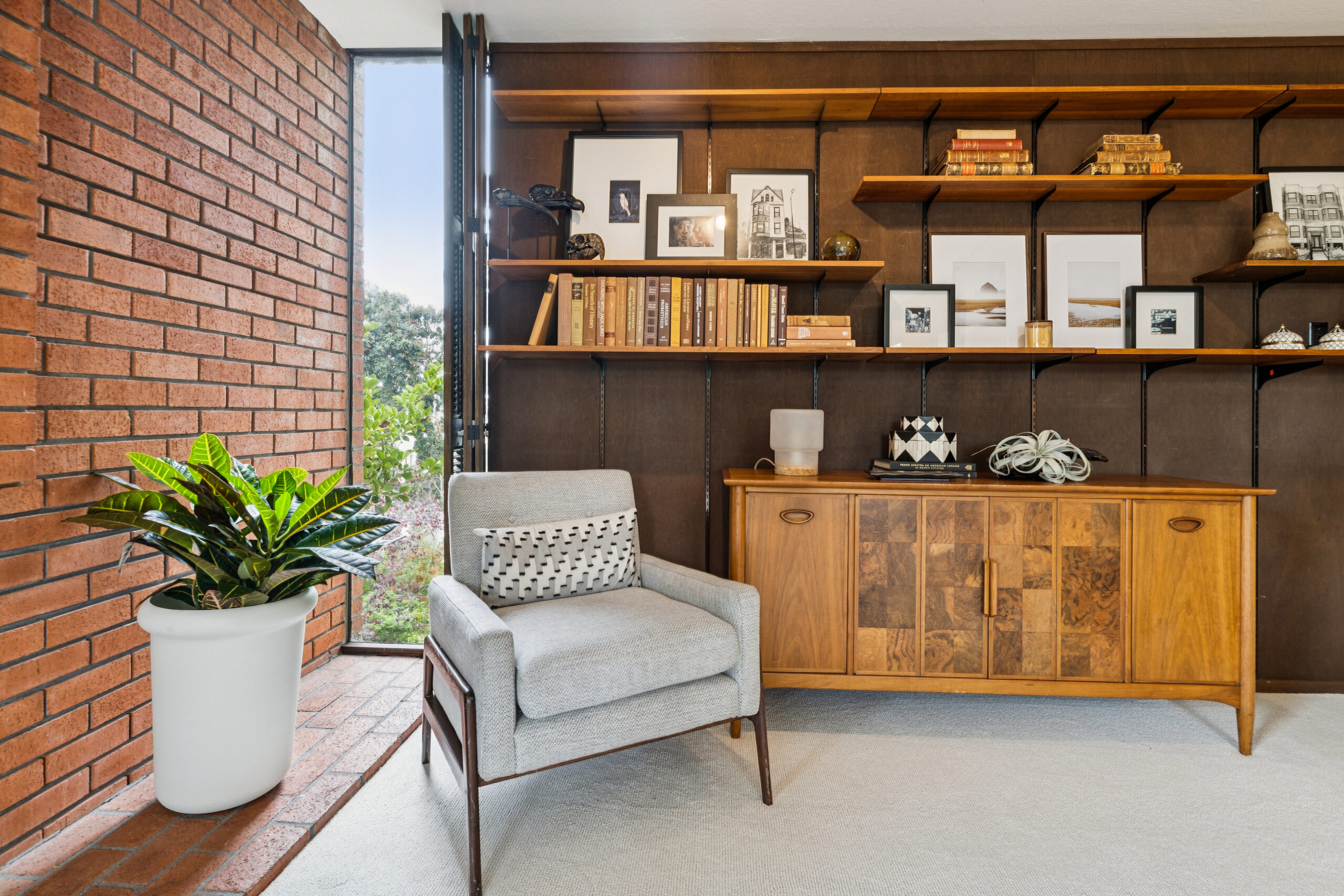 45 Cleary Court A gray armchair with a patterned lumbar pillow sits in front of wall-mounted shelving and a wooden sideboard, next to a large potted plant and a brick wall.