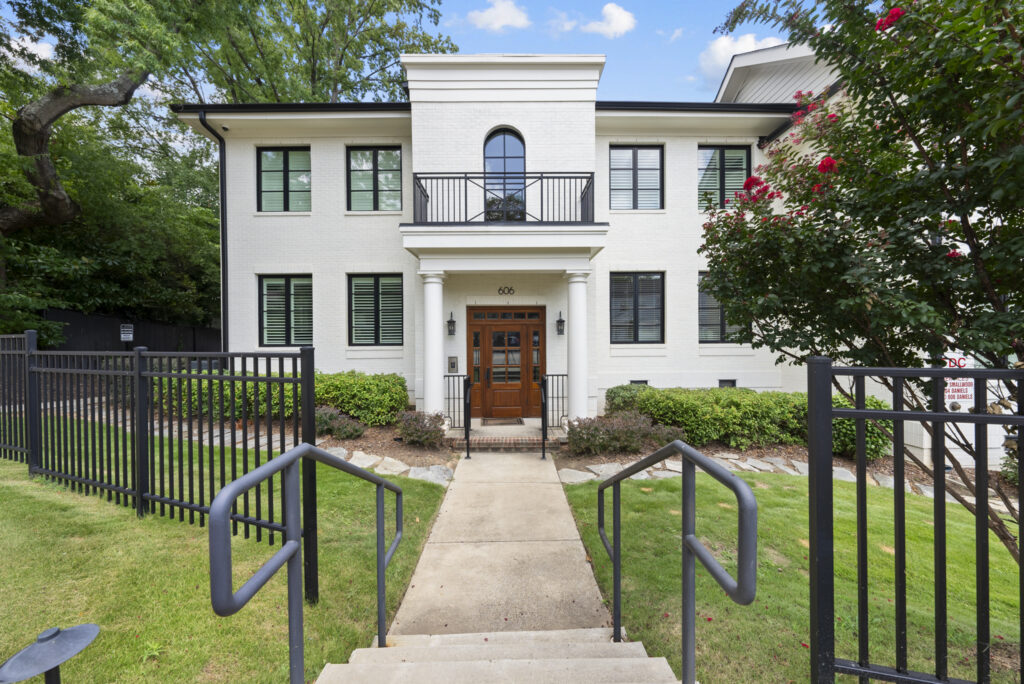 A two-story white brick building with black window shutters, a wooden front door, a small balcony, and a walkway leading up to the entrance, nestled in a college town and surrounded by fenced green grass and trees.