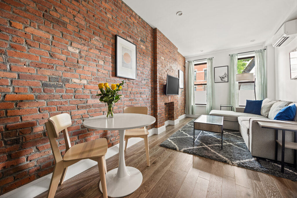 A living and dining area featuring exposed brick walls, a round white table with two wooden chairs, a vase of yellow flowers, a gray sofa, wall-mounted TV, large windows with sheer curtains, and wooden floors.