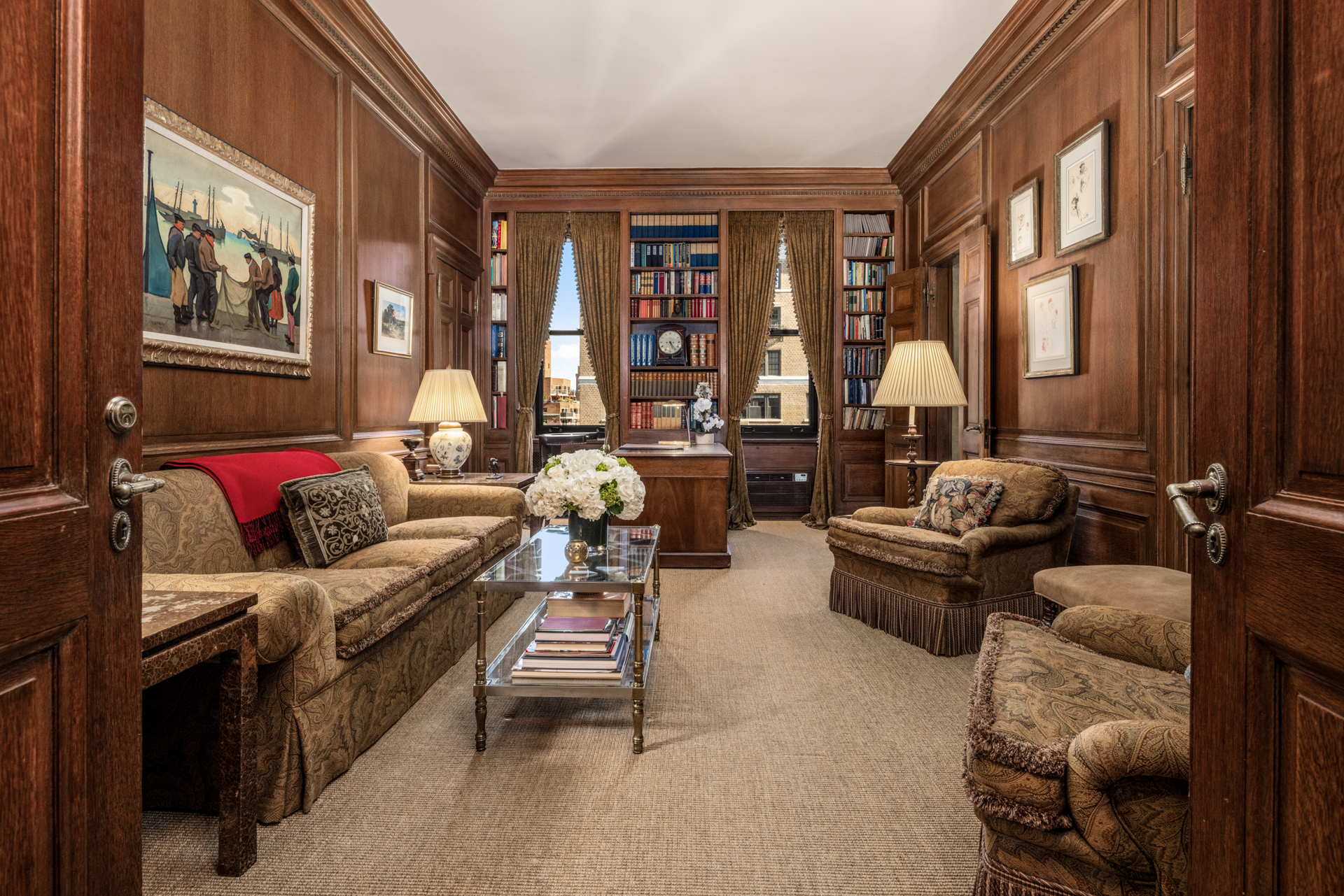 812 Park Avenue, 11 and 12B A wood-paneled home office library featuring a sofa with a red throw, two armchairs, a coffee table with flowers, and floor-to-ceiling bookshelves flanking a window.