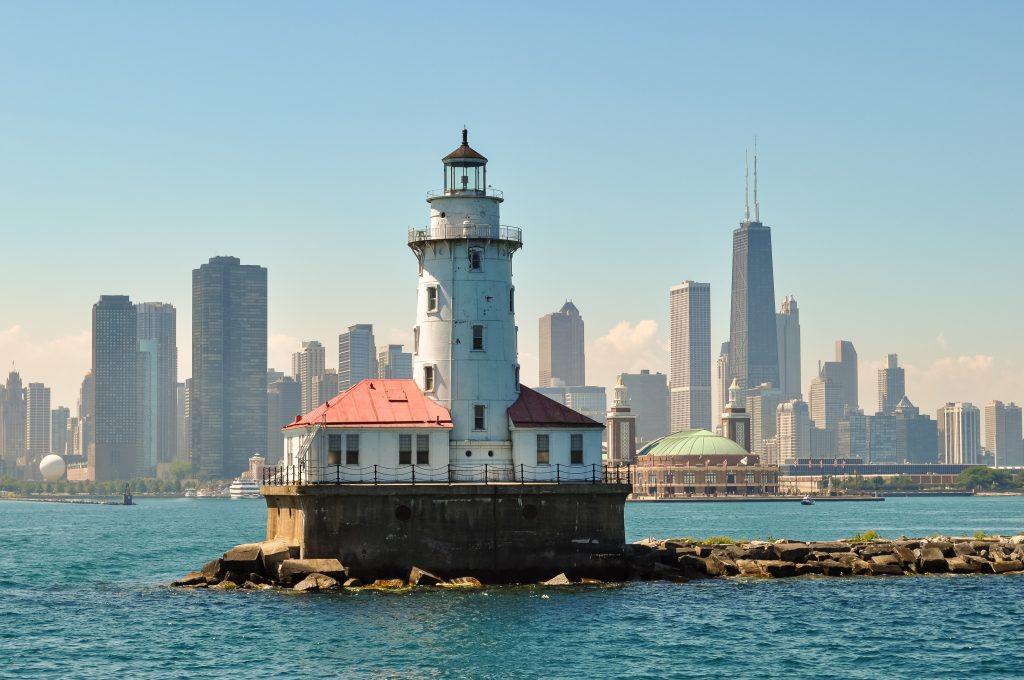 The Chicago Harbor Lighthouse sits on a breakwater in Lake Michigan on a sunny day with the city skyline in the background.