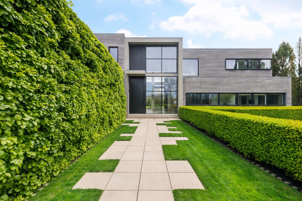 Looking straight-on at 183 Daniels Lane in Sagaponack, up the asymmetrical front walkway. A wall of glass reveals the interior's floating travertine staircase.