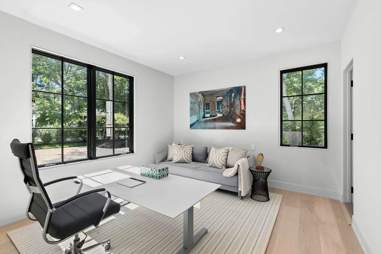 Modern home offices shine in this space with large windows, a sleek white desk, black office chair, gray sofa with throw pillows, side table, and wall art. Natural light fills the room with views of greenery outside.