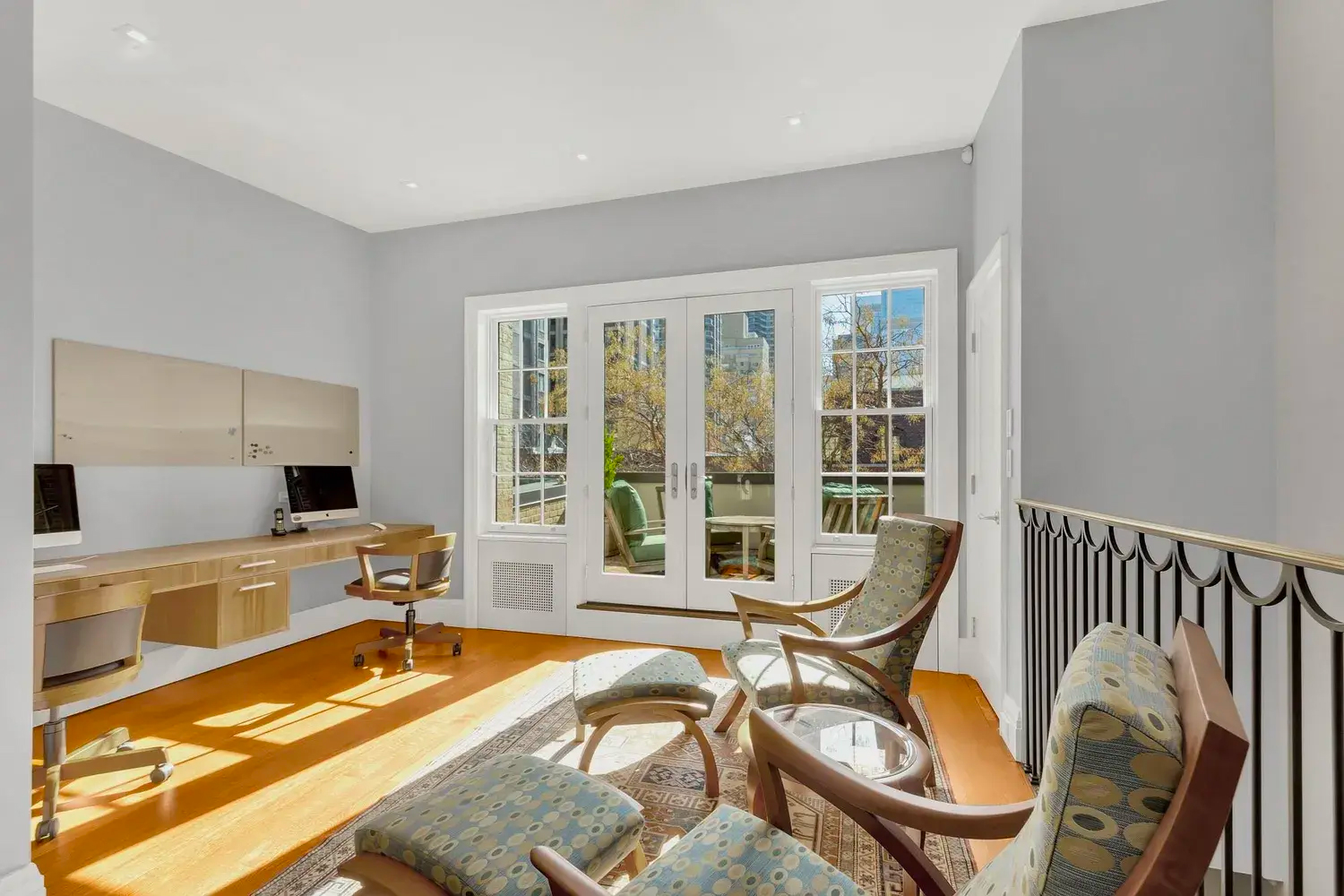 A bright home office with two modern desks and computers, several patterned chairs, and large glass doors opening to a garden—an inspiring example of stylish home offices. Sunlight streams in, illuminating the wooden floor and light gray walls.