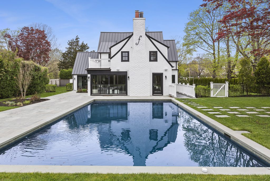 A white house with a chimney sits at the head of a vibrantly pool swimming pool in Southampton.