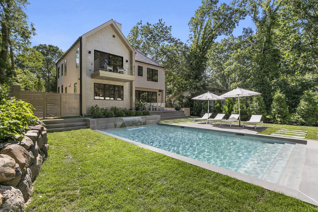 The verdant backyard at 40 Lincoln Street in Sag Harbor boasts a heated swimming pool with a miniature waterfall, a patio, and a covered fireplace lounge.