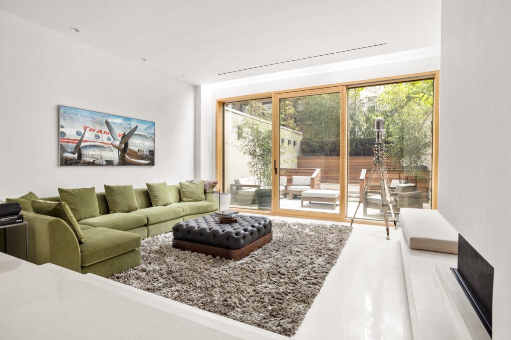 A sunken living room looks out onto a back garden through tall glass doors. The room sports a green couch, black leather ottoman, and a gas fireplace.