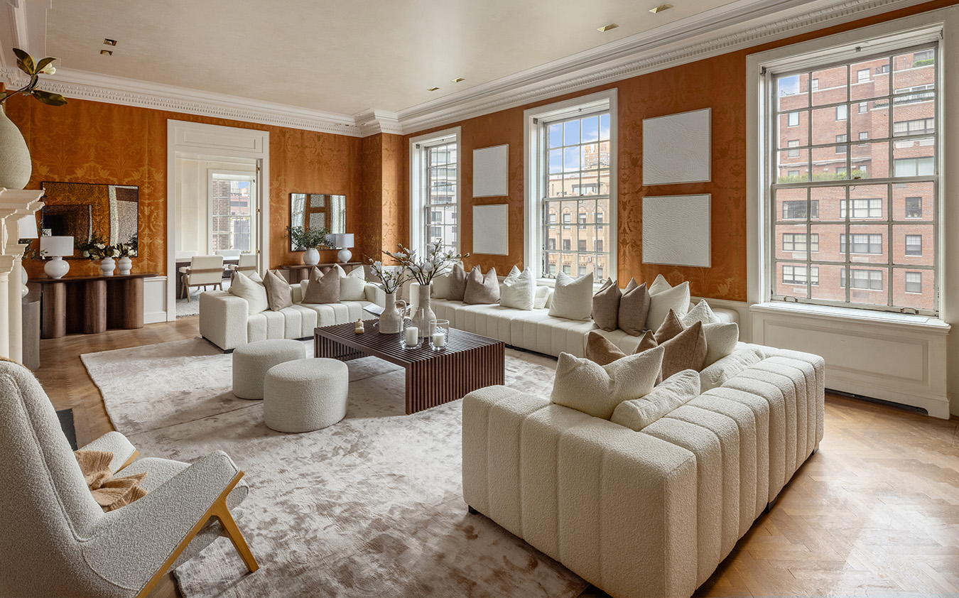A luxurious living room with large windows, white sofas, plush pillows, a wooden coffee table, soft beige rug, and warm orange walls, with city buildings visible outside. Modern art and decor accent the spacious, elegant space.