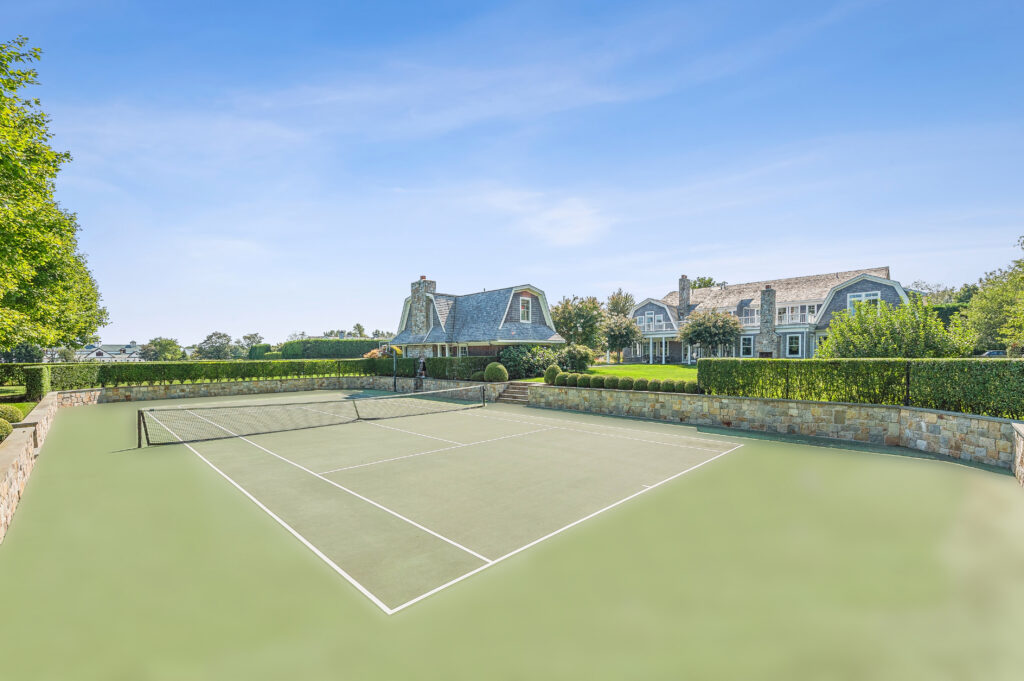 A well-maintained outdoor tennis court with a net, one of the desirable summer amenities, is surrounded by lush greenery and stone walls, set in front of two large, elegant houses under a clear blue sky.
