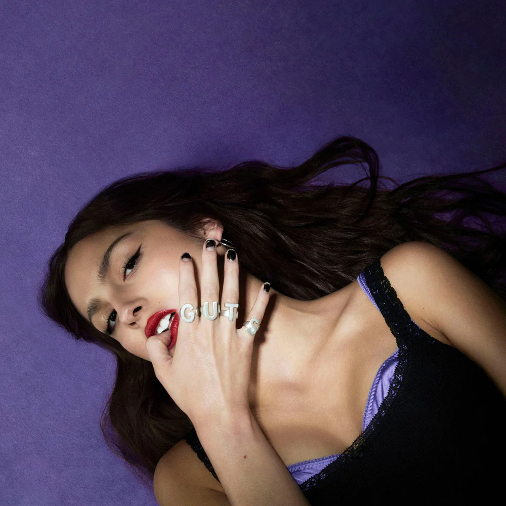 Olivia Rodrigo lays against a purple background with her thumb in between her teeth and the album's title GUTS spelled out on individual rings on her other four fingers
