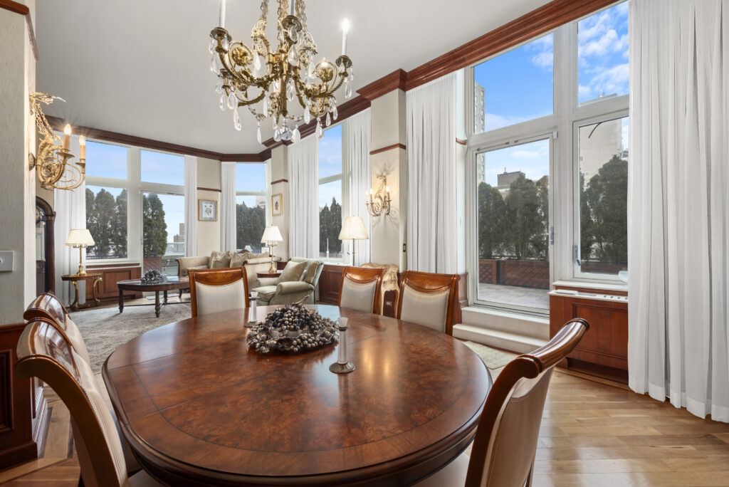 An elegant living and dining room sat atop diagonal hardwood flooring. The walls and ceilings are white with brown wooden moldings and accents. There is an entrance to a wraparound terrace from two points in the room.