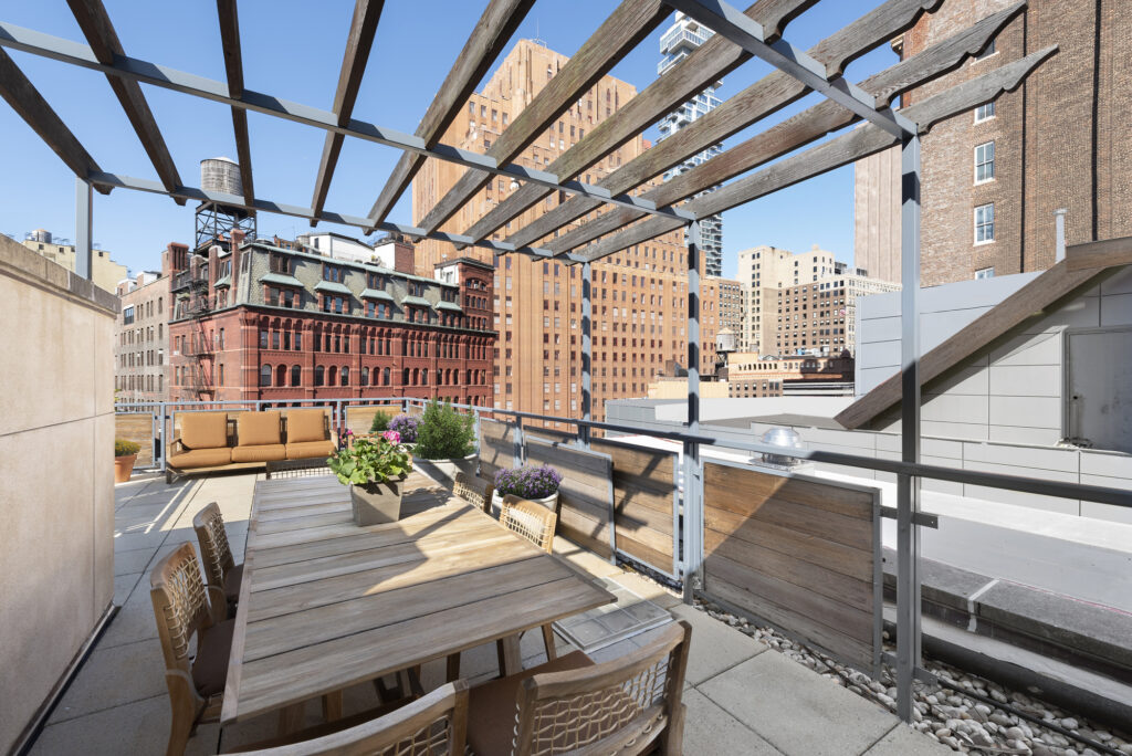 A pergola sits above an outdoor dining table, surrounded by chairs with wicker backrests. In the distance sits a brownish-orange three-seat couch with views of iconic Tribeca architecture.