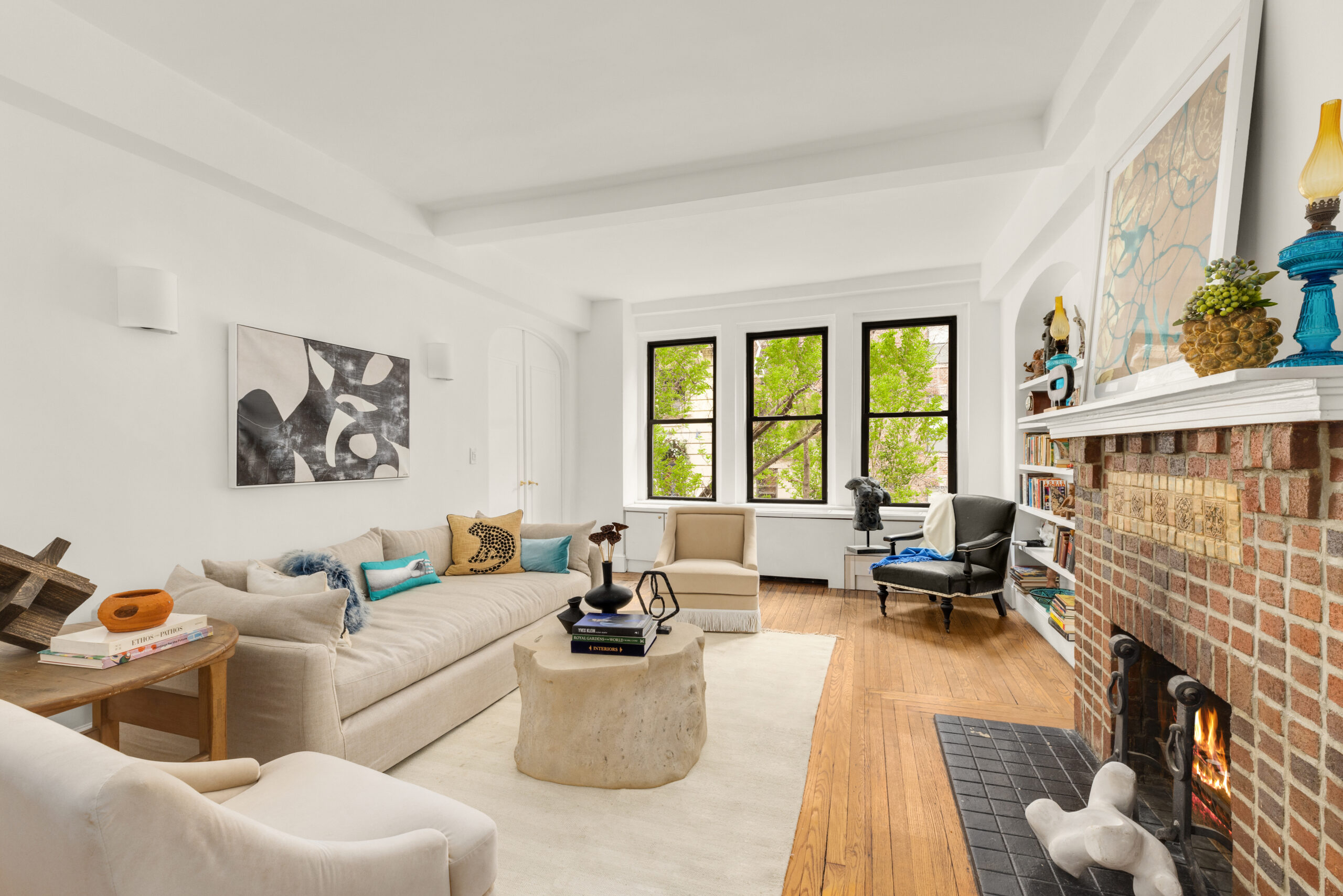 Bright living room with large windows, white walls, beige sofas, hardwood floors, and a modern abstract painting. A stylish brick fireplace decorated with colorful objects adds warmth to the space. A tree is visible outside the windows.