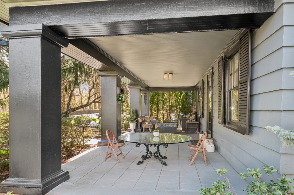 11 Midland Avenue, Bronxville A spacious covered front porch, featuring gray siding, black columns, a round glass table with chairs, potted plants, and outdoor seating set—perfect for those who love stylish front porches surrounded by lush greenery.