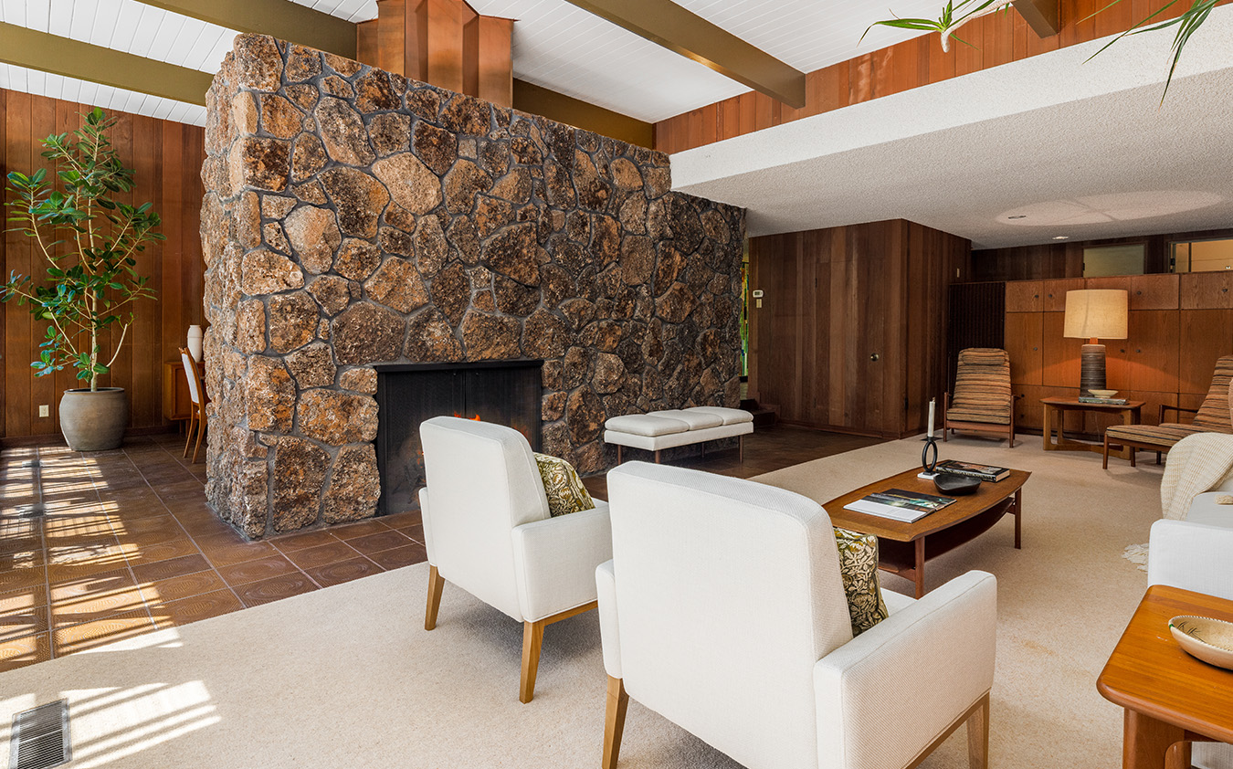 A mid-century modern living room with a large stone fireplace, white armchairs, a wooden coffee table, and wood-paneled walls. Sunlight streams in from windows, and a tall plant stands in the corner.