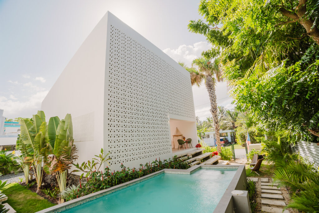 A modern white house with a perforated facade stands next to a rectangular swimming pool, surrounded by lush tropical plants and palm trees—perfectly capturing the vibrant energy of aries season under a bright, sunny sky.