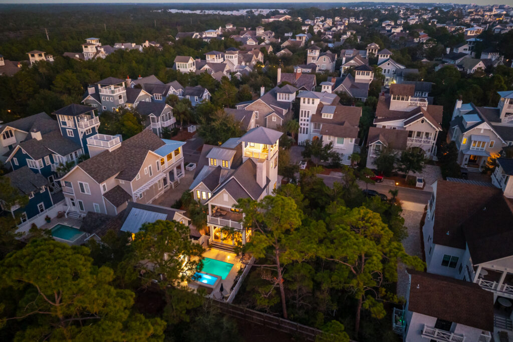 Aerial view of a residential neighborhood at dusk during aries season, featuring modern multi-story houses surrounded by trees, with a brightly lit swimming pool in the backyard of one home.