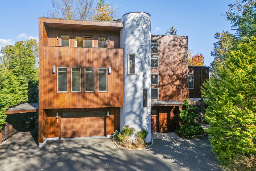 Modern, multi-story wooden house with large windows and a prominent cylindrical white tower in the center, surrounded by lush green trees basking in bright sunlight.