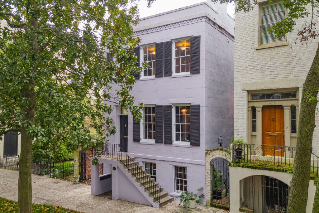 A two-story light purple townhouse with black shutters, large windows, and an exterior staircase leading to a black front door, surrounded by trees and greenery—perfect for embracing new beginnings during Aries season.