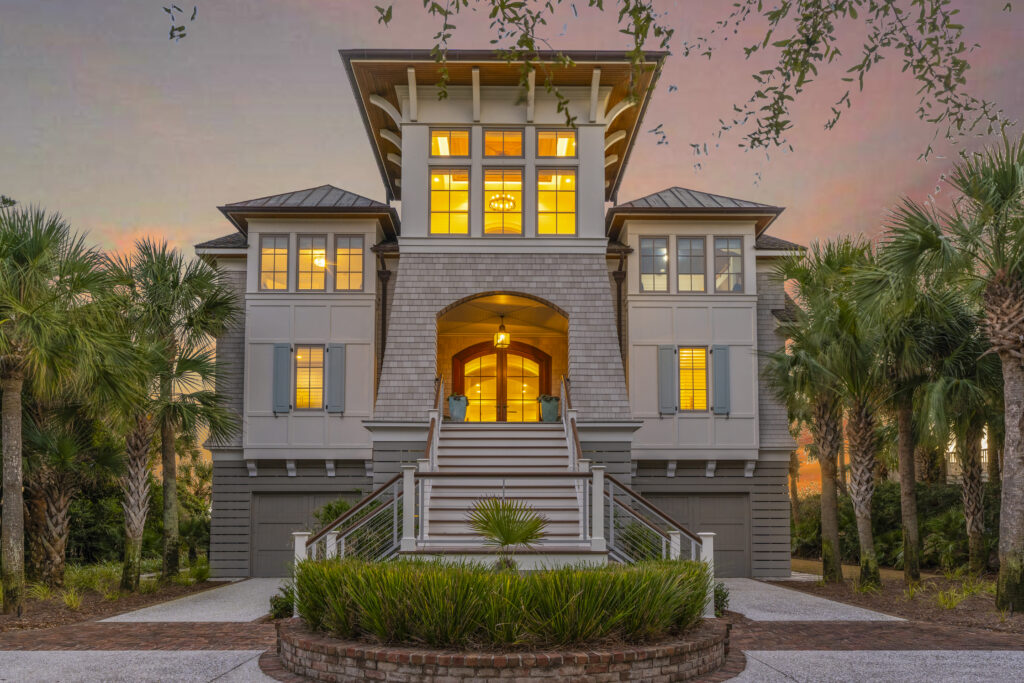 A large, elegant house with many windows and warm interior lighting, surrounded by palm trees, features a grand staircase leading to the entrance at sunset—perfectly capturing the vibrant energy of Aries season.