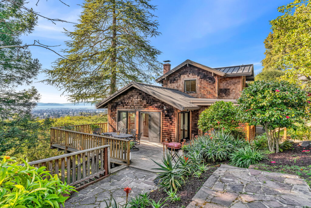 A rustic two-story house with wooden siding, surrounded by lush greenery, a stone patio, outdoor seating, and a scenic view of the city and water—perfect for soaking in the fresh energy of Aries season under a clear blue sky.