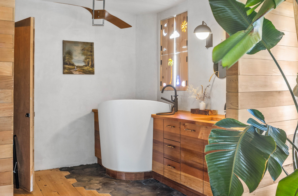 A deep white soaking bathtub sits in front of custom wood shutters inlaid with glass shapes.