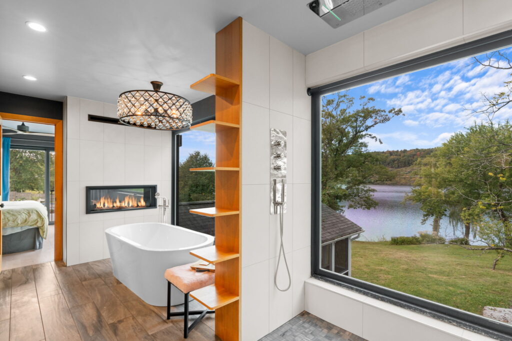 A bathtub sits beneath a chandelier, beside a fireplace, and looks out toward a lake in the distance.