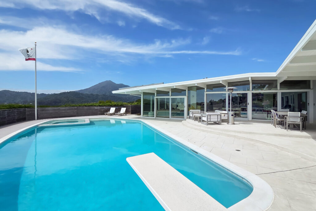 Modern house with large windows and white patio furniture surrounds a clear blue swimming pool. A flag waves on the left, with a mountain and blue sky in the background—capturing that relaxed, sunny Gemini season vibe outdoors.