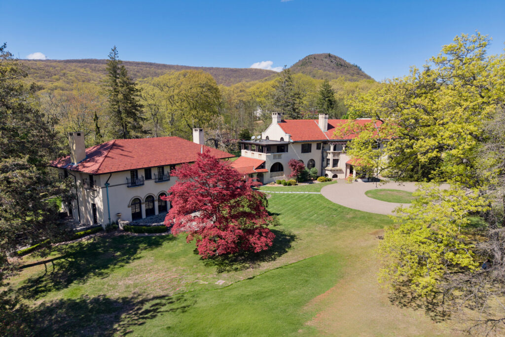 A large mansion with red-tiled roofs and cream-colored walls sits amid lush green lawns, flowering trees, and surrounding woods—perfectly capturing the vibrant energy of Gemini season—with hills rising in the background under a clear blue sky.