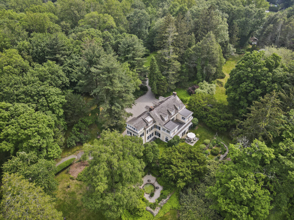 A large, elegant house surrounded by dense green trees and landscaped gardens, viewed from above. Embrace peaceful vibes this Gemini season with a circular driveway, patio, and well-maintained lawns in a serene wooded area.