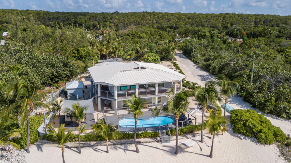 Aerial view of a modern beachfront house with a pool, surrounded by palm trees and greenery, basking in the vibrant energy of Gemini season on white sand near a forested area.