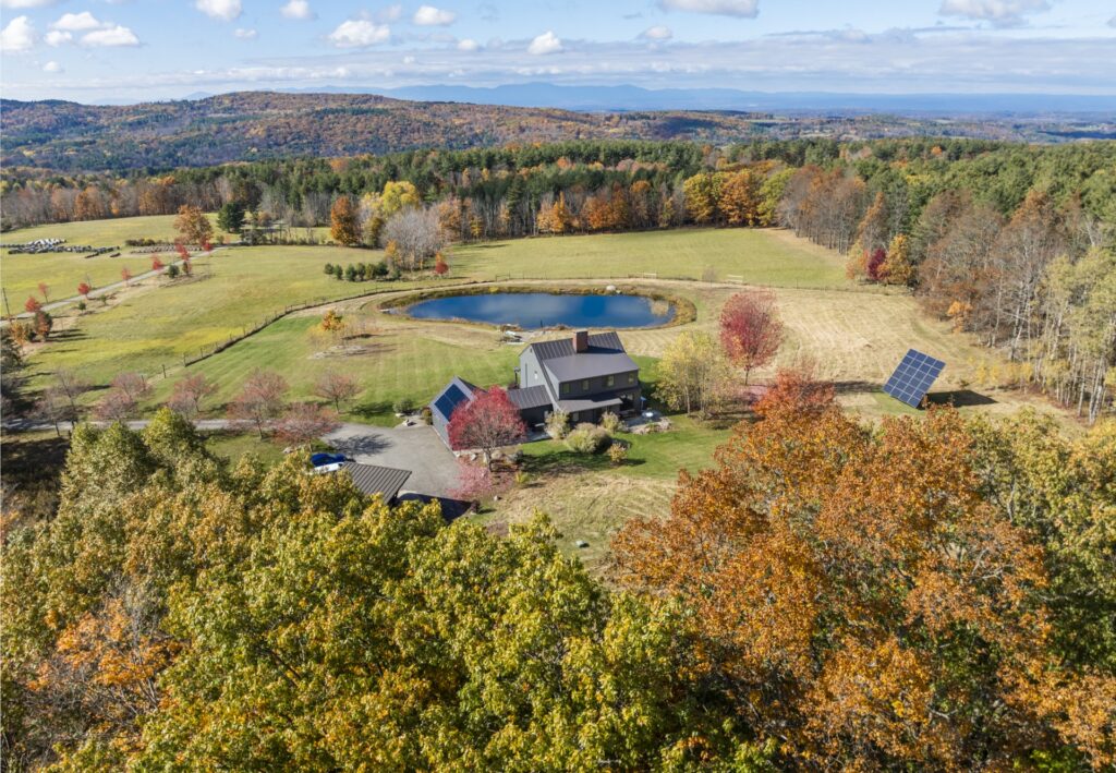 Aerial view of a rural house surrounded by autumn trees, open fields, and solar panels basking in the light of gemini season, with rolling hills, a pond, and distant mountains under a partly cloudy sky.