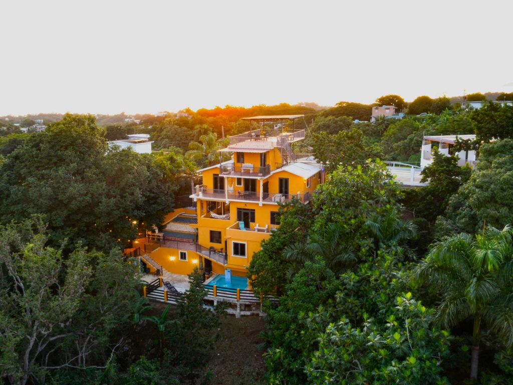 A vibrant yellow multi-story house with balconies sits among lush green trees at sunset, surrounded by dense vegetation and a few neighboring buildings, radiating the lively energy of Gemini season.