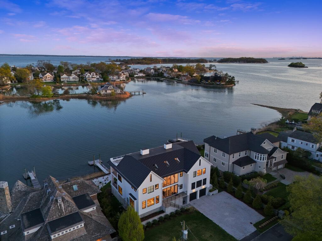 Aerial view of large waterfront homes along a calm bay at sunset during gemini season, with reflections on the water and a vibrant sky in shades of pink and blue.