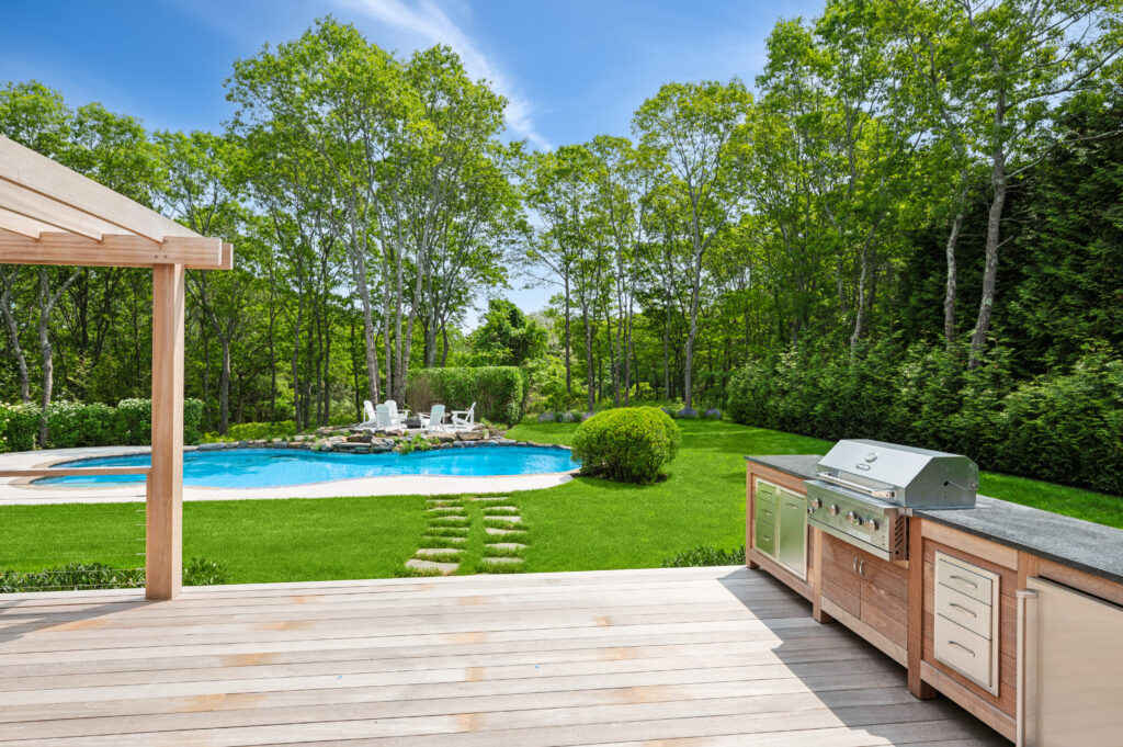 Outdoor kitchen area with a built-in grill and countertop on a wooden deck overlooking a lush green backyard with a swimming pool and lounge chairs.