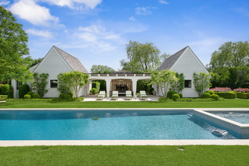 A luxurious backyard features a large swimming pool with a hot tub, surrounded by lush green grass and trees, leading to a white house with two peaked roofs and an outdoor seating area under a pergola.