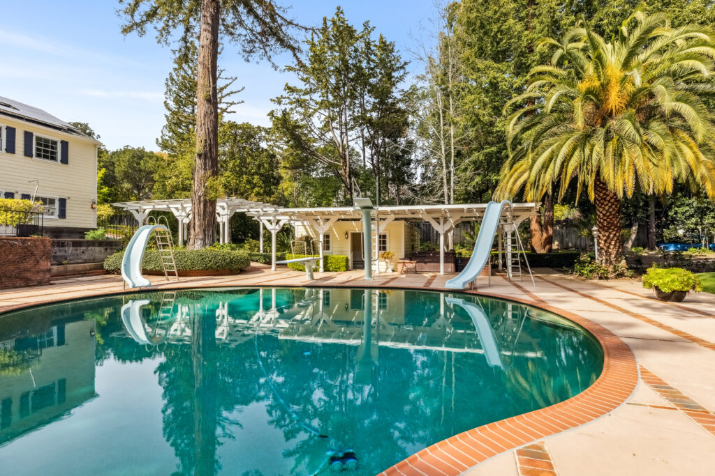 Curved outdoor swimming pool with two slides, surrounded by a tiled deck, landscaped trees and plants, a pergola with seating, and a yellow house with white trim on the left.
