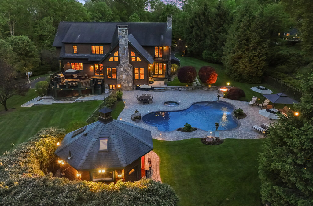 Large multi-story luxury house with a steep roof, illuminated windows, an irregularly shaped swimming pool, patio furniture, a lit pool house, and lush landscaping surrounded by dense trees at night.
