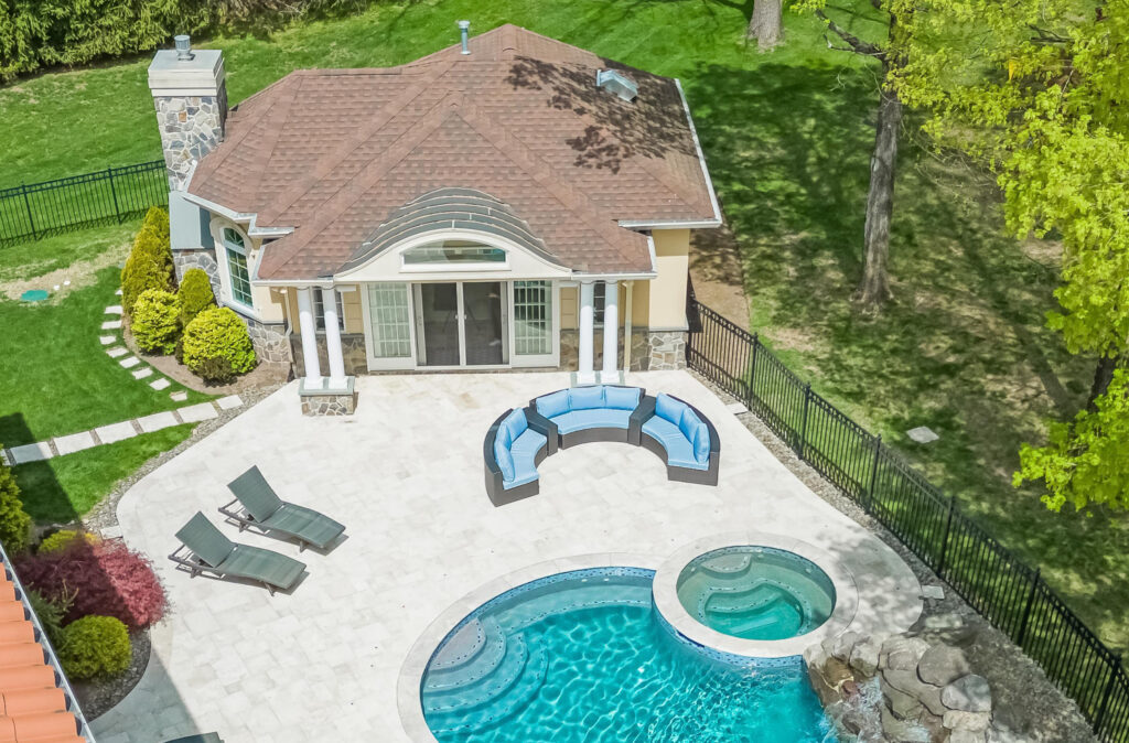 Aerial view of a backyard with a beige pool house with a brown roof, a swimming pool with an attached hot tub, curved sectional patio sofa, lounge chairs, and surrounding greenery and trees.